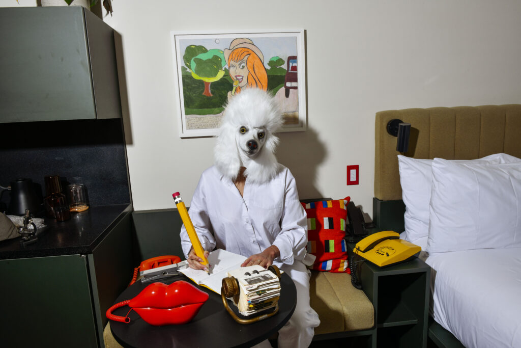 A man wearing a Dogs headpiece in a Hotel Room, seated, taking notes by the bed with Art decoration