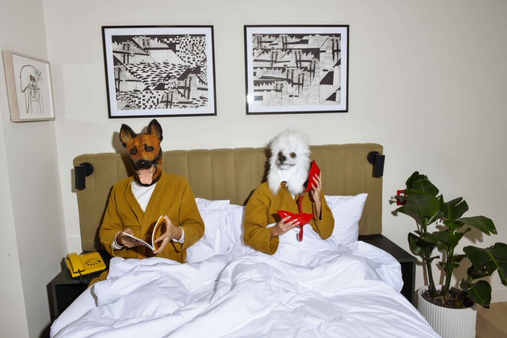Two adults wearing dogs masks laying in a bed, one is reading a magazine and the other one with a red phone